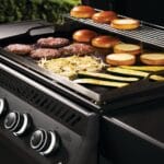 Grill with burgers, buns, and vegetables.